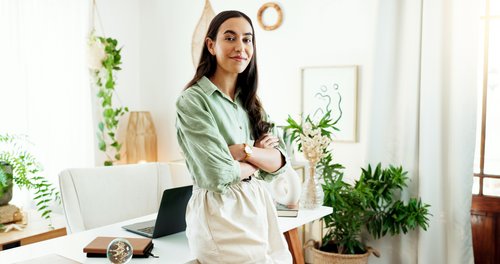 A woman with long, dark hair stands in a bright, modern office with plants and a laptop on the table.