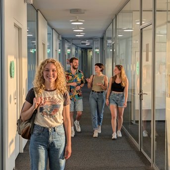 A group of four people walks down a modern hallway with glass walls. A woman with curly hair smiles at the camera.
