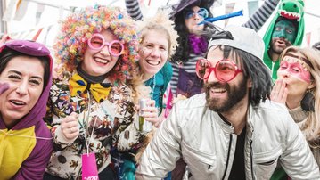 A group of people in colorful costumes and accessories, joyfully posing and celebrating. Some are wearing hats and oversized sunglasses.