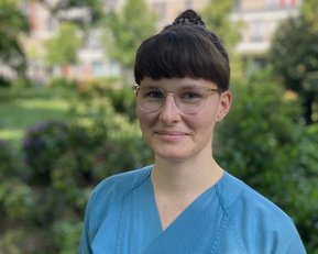 A person with short brown hair and wearing blue work clothes stands outdoors, surrounded by plants and trees.