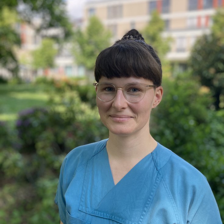 A person with short brown hair and wearing blue work clothes stands outdoors, surrounded by plants and trees.
