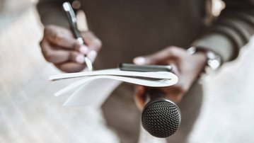 Eine Hand hält ein Mikrofon, während die andere auf einem Notizblock Notizen macht.