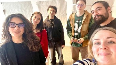 Six people pose for a group photo in a bright room, with drawings in the background. Everyone is smiling at the camera.