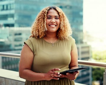 A woman with curly hair is holding a tablet and smiling while standing on a terrace overlooking a city.