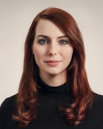 A woman with long red hair and blue eyes is wearing a black top. She is looking directly at the camera.