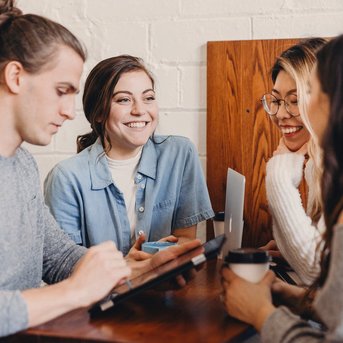 A group of four young people is sitting at a table in a café, smiling and chatting, while one of them is typing on a tablet.