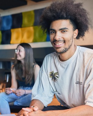 Ein junger Mann mit lockigem Haar sitzt lächelnd im Vordergrund, während zwei Frauen im Hintergrund miteinander sprechen. Alle drei sind in einem modernen Raum mit bunten Wanddekorationen.