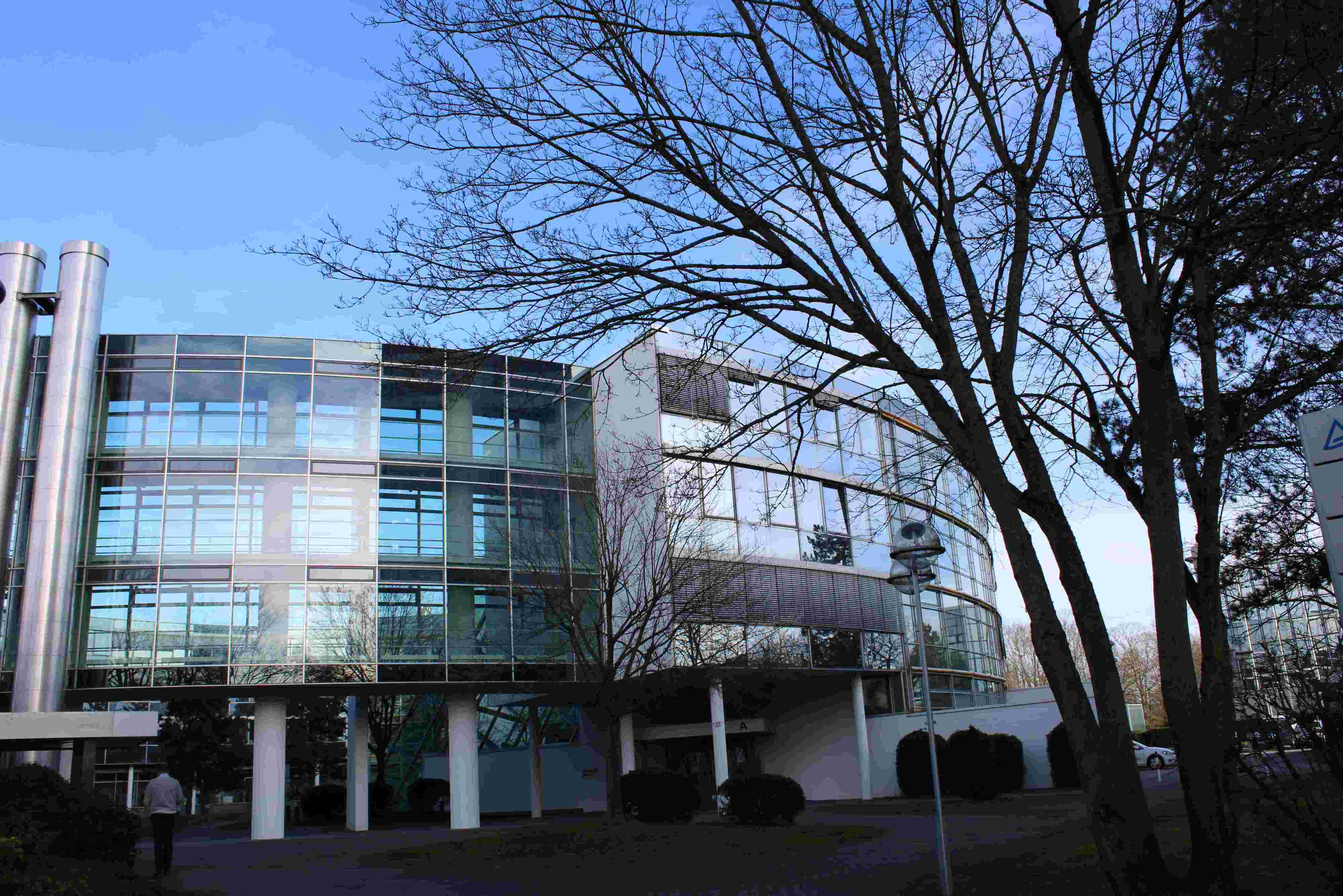Moderne Bürogebäude mit großen Fenstern und einem klaren, minimalistischen Design, umgeben von Bäumen und einem Gehweg. Der Himmel ist blau und klar.