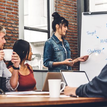 Eine Frau präsentiert einen Marketingplan an einem Flipchart, während drei Personen am Tisch aufmerksam zuhören. Der Raum hat eine moderne, industrielle Atmosphäre mit Ziegelwänden und großen Fenstern.