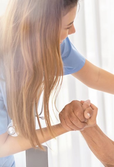 Eine Pflegekraft hält die Hand eines lächelnden älteren Mannes, der in einem Rollstuhl sitzt, in einem hellen Raum.