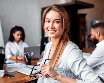 Eine lächelnde Frau hält eine Brille in einem Besprechungsraum, während drei Personen am Tisch mit Laptops und Notizen arbeiten.