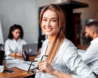 A smiling woman holds a pair of glasses in a meeting room while three people work at the table.