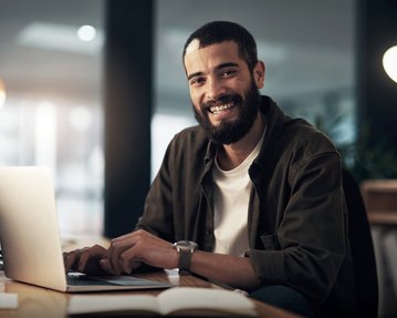 Ein Mann mit Bart sitzt an einem Schreibtisch und lächelt, während er an einem Laptop arbeitet. Helle Büroumgebung im Hintergrund.