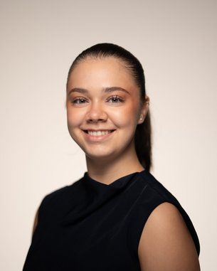 Eine junge Frau mit einem freundlichen Lächeln trägt ein schwarzes Oberteil und hat ihre Haare zu einem Pferdeschwanz gebunden.