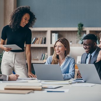 Eine Gruppe von fünf Personen in einem modernen Büro, die an einem Tisch sitzen. Eine Frau steht und spricht, während die anderen lachen und aufmerksam zuhören. Laptops und Notizen sind auf dem Tisch verteilt.