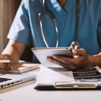 Ein Arzt in blauer Arbeitskleidung nutzt ein Tablet, während er an einem Laptop arbeitet. Auf dem Tisch liegen Notizblätter und ein Stethoskop.