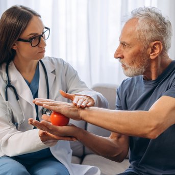 Eine Ärztin untersucht die Hand eines älteren Mannes, während sie ihm eine orangefarbene Kugel zeigt. Beide sitzen auf einem Sofa.