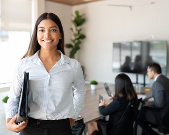 Eine junge Frau in einem Büro trägt eine Bluse und hält eine Mappe. Im Hintergrund sind zwei Personen an einem Tisch mit Laptops zu sehen.