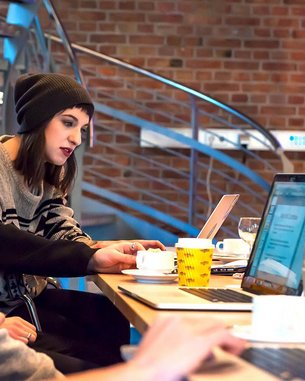 A group of four people is sitting at a table with laptops. One person in a black hat is interacting with a laptop while the others watch attentively. On the table, there are cups and a yellow mug.