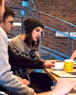 Eine Gruppe von vier Personen sitzt an einem Tisch mit Laptops. Eine Person in einem schwarzen Hut interagiert mit einem Laptop, während die anderen aufmerksam zuschauen. Auf dem Tisch stehen Tassen und ein gelbes Becher.