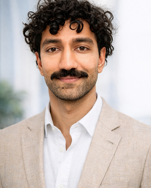 Ein Mann mit lockigem Haar und einem Schnurrbart trägt einen hellen Anzug und ein weißes Hemd. Der Hintergrund ist unscharf.