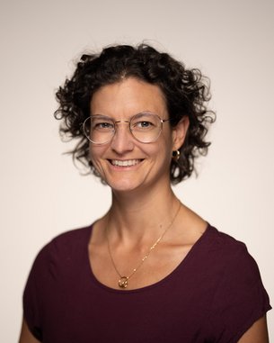 Eine Frau mit lockigem Haar und Brille lächelt in die Kamera, trägt ein lila Oberteil und hat eine goldene Halskette an.