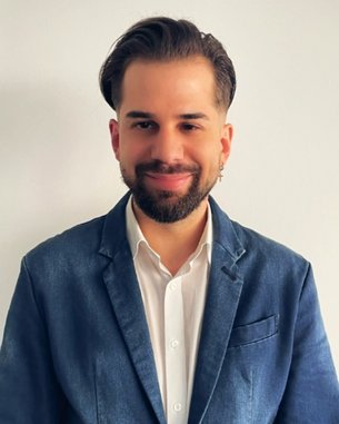 Mann mit Bart und Ohrring trägt einen blauen Blazer über einem weißen Hemd, steht vor einer neutralen Wand.