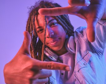 A young man with dreadlocks poses with both hands, simulating a camera perspective, against a purple background.