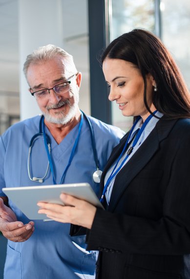 A doctor and a businesswoman are standing in a modern office, looking at a tablet together. Both are smiling and seem to be discussing the information on the screen.