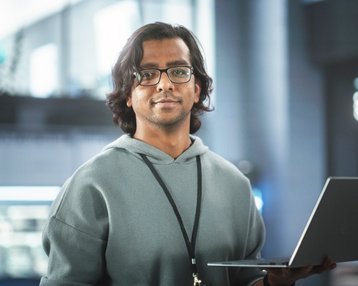 Ein junger Mann mit Brille hält einen Laptop in einem modernen, hellen Raum. Er trägt einen grauen Hoodie und schaut direkt in die Kamera.