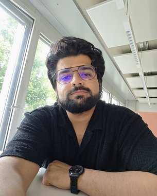 Ein Mann mit dunklem, lockigem Haar und einer Brille sitzt an einem Tisch in einem hellen Raum mit Fenstern.