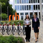 Zwei Frauen gehen lächelnd auf einem gepflasterten Weg vorbei an einer Reihe von Fahrrädern, mit einem modernen Gebäude im Hintergrund und grünen Pflanzen um sie herum.