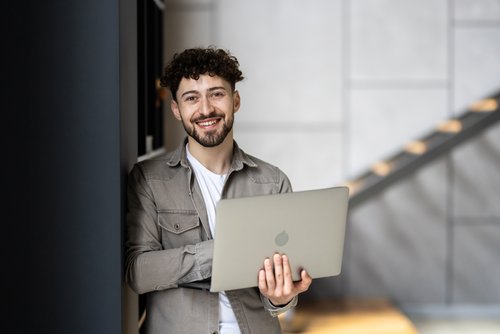 Ein junger Mann mit lockigem Haar steht an einer Wand und hält einen Laptop in den Händen, während er lächelt.