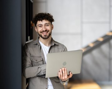A young man with curly hair stands by a wall, holding a laptop in his hands while smiling.