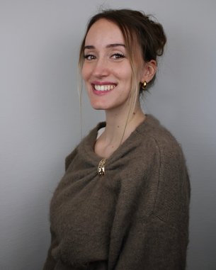 Eine lächelnde Frau mit langen, lockigen Haaren trägt einen beigen Pullover und goldene Ohrringe. Der Hintergrund ist neutral.