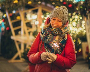 A woman in a red coat and gray hat holds a sparkler, surrounded by festive lights and decorations.