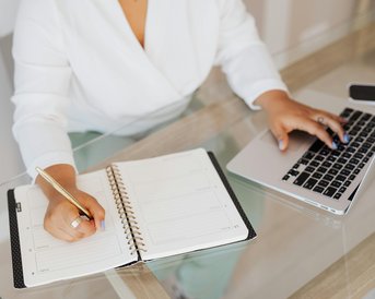 Eine Person schreibt in ein Notizbuch, während sie an einem Laptop arbeitet. Der Tisch ist aus Glas und die Umgebung ist hell und modern.