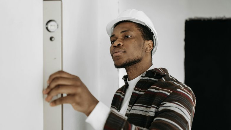 Ein Mann mit einem weißen Helm überprüft mit einer Wasserwaage die Ebenheit einer Wand. Er trägt ein gestreiftes Hemd und konzentriert sich auf seine Aufgabe.