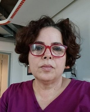 Frau mit lockigem, braunem Haar und roten Brillen sitzt in einem modernen Raum, trägt ein lila Oberteil und schaut direkt in die Kamera.