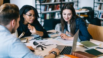 Drei Studierende arbeiten an einem Tisch, umgeben von Notizen und Laptops, während sie gemeinsam an einem Projekt diskutieren.