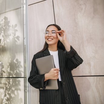 A smiling woman in a striped suit holds a laptop and looks through her glasses. She stands in front of a modern glass wall that reflects the light and the plants.