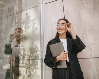 A smiling woman in a striped suit holds a laptop and looks through her glasses. She stands in front of a modern glass wall that reflects the light and the plants.