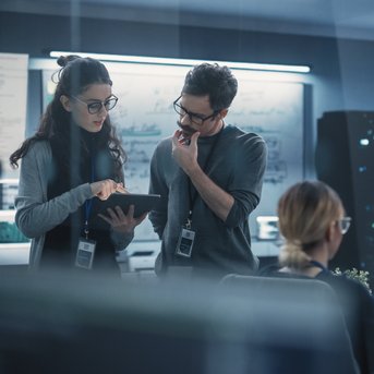 Two people in a modern office are discussing something on a tablet, while another person is working at a computer.