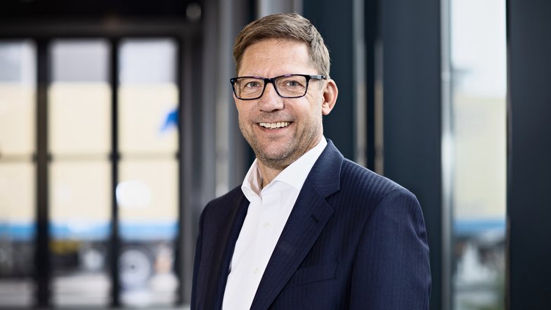 Ein Mann mit Brille und Anzug lächelt vor einer modernen Glaswand. Er hat kurzes, braunes Haar und wirkt professionell.