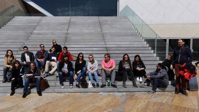 Eine Gruppe von etwa 20 Personen sitzt auf einer Treppe vor einem modernen Gebäude. Die Teilnehmer sind in verschiedenen Positionen angeordnet und tragen legere Kleidung. Einige halten persönliche Gegenstände oder Notizen in den Händen.