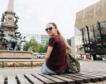 Young woman with sunglasses sitting on a bench, with a fountain and modern buildings in the background.