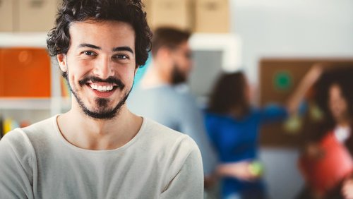 Lächelnder Mann mit dunklen, lockigen Haaren steht im Vordergrund, während Menschen im Hintergrund miteinander interagieren.