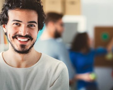 Lächelnder Mann mit dunklen, lockigen Haaren steht im Vordergrund, während Menschen im Hintergrund miteinander interagieren.