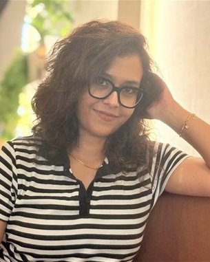 Eine Person mit lockigem Haar und Brille sitzt auf einem Sofa und lächelt, während sie eine Hand hinter den Kopf legt. Sie trägt ein gestreiftes Oberteil.