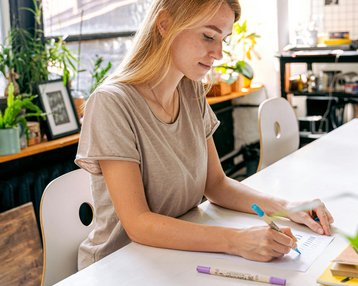 Eine junge Frau sitzt an einem Tisch in einem kreativen Raum und zeichnet mit Stiften auf Papier. Um sie herum sind Pflanzen und verschiedene Kunstmaterialien sichtbar.